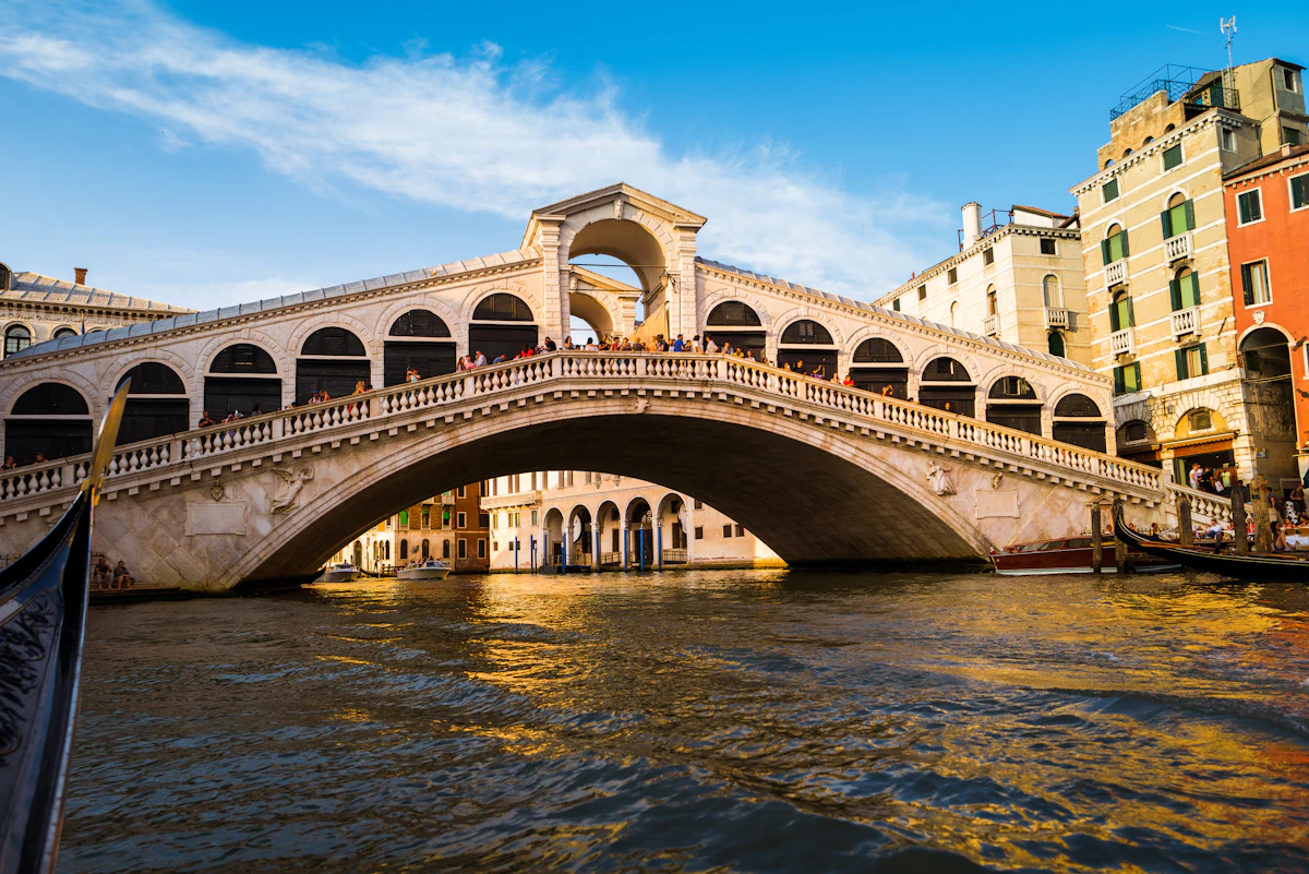 Die weiße Steinbrücke Rialto, die sich bei Tageslicht über den Canal Grande in Venedig wölbt, mit einer darunter hindurchfahrenden Gondel und orangefarbenen Gebäuden am Wasser