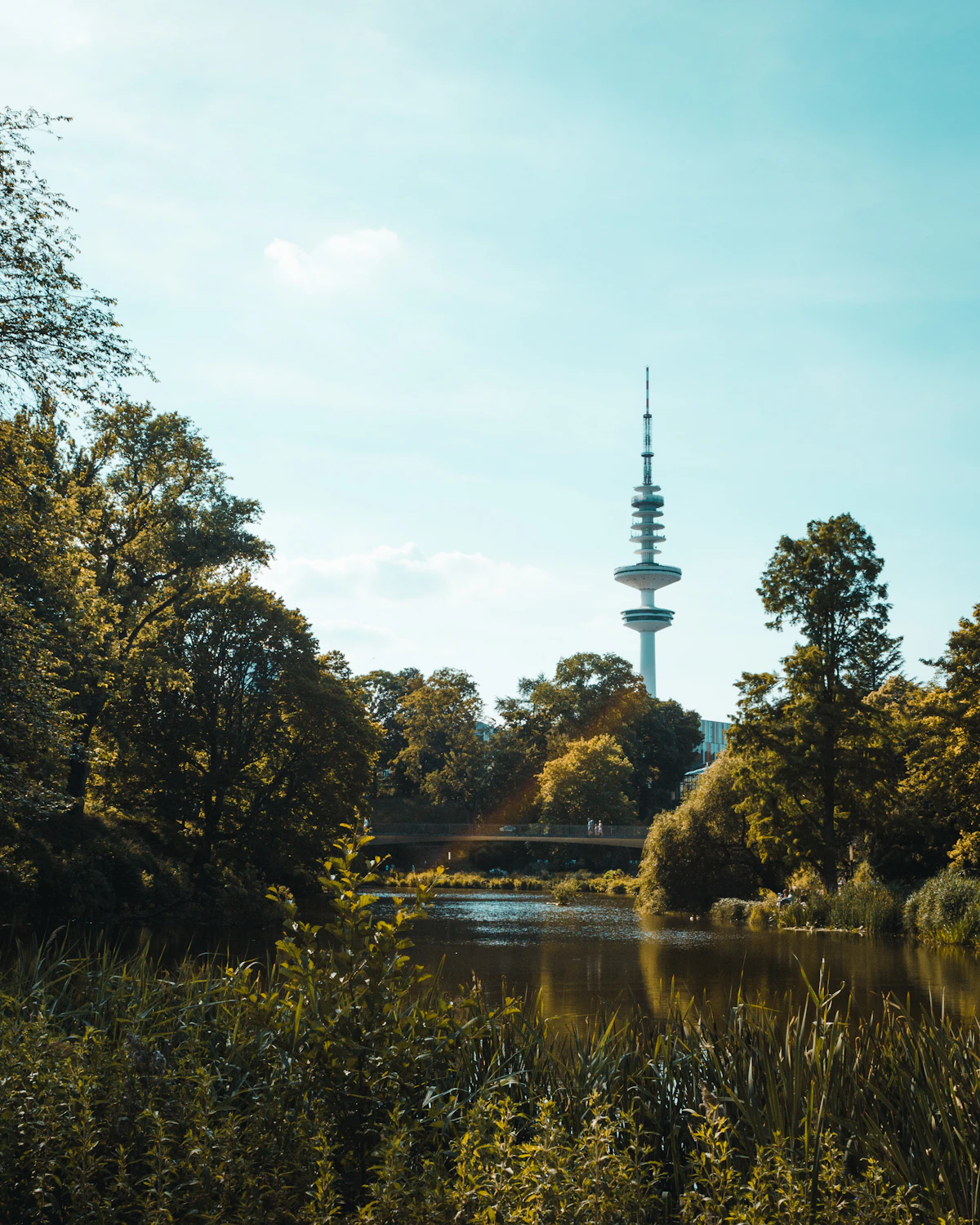 Um lago no parque Planten un Blomen emoldurado por árvores outonais, com a torre de televisão Heinrich-Hertz-Turm de Hamburgo visível por detrás da linha das árvores