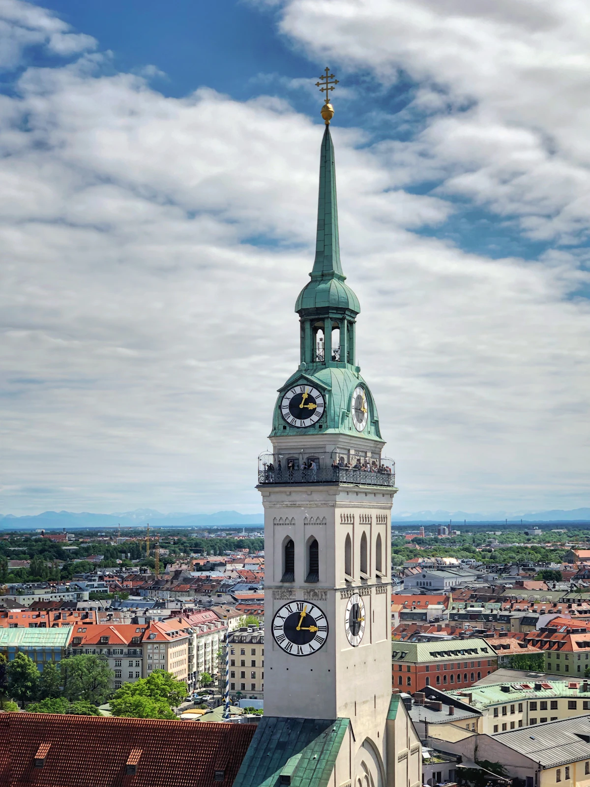 Der Uhrturm mit grüner Spitze der Kirche St. Peter (Alter Peter) aus der Nähe, mit Münchner Dächern und den Alpen in der Ferne