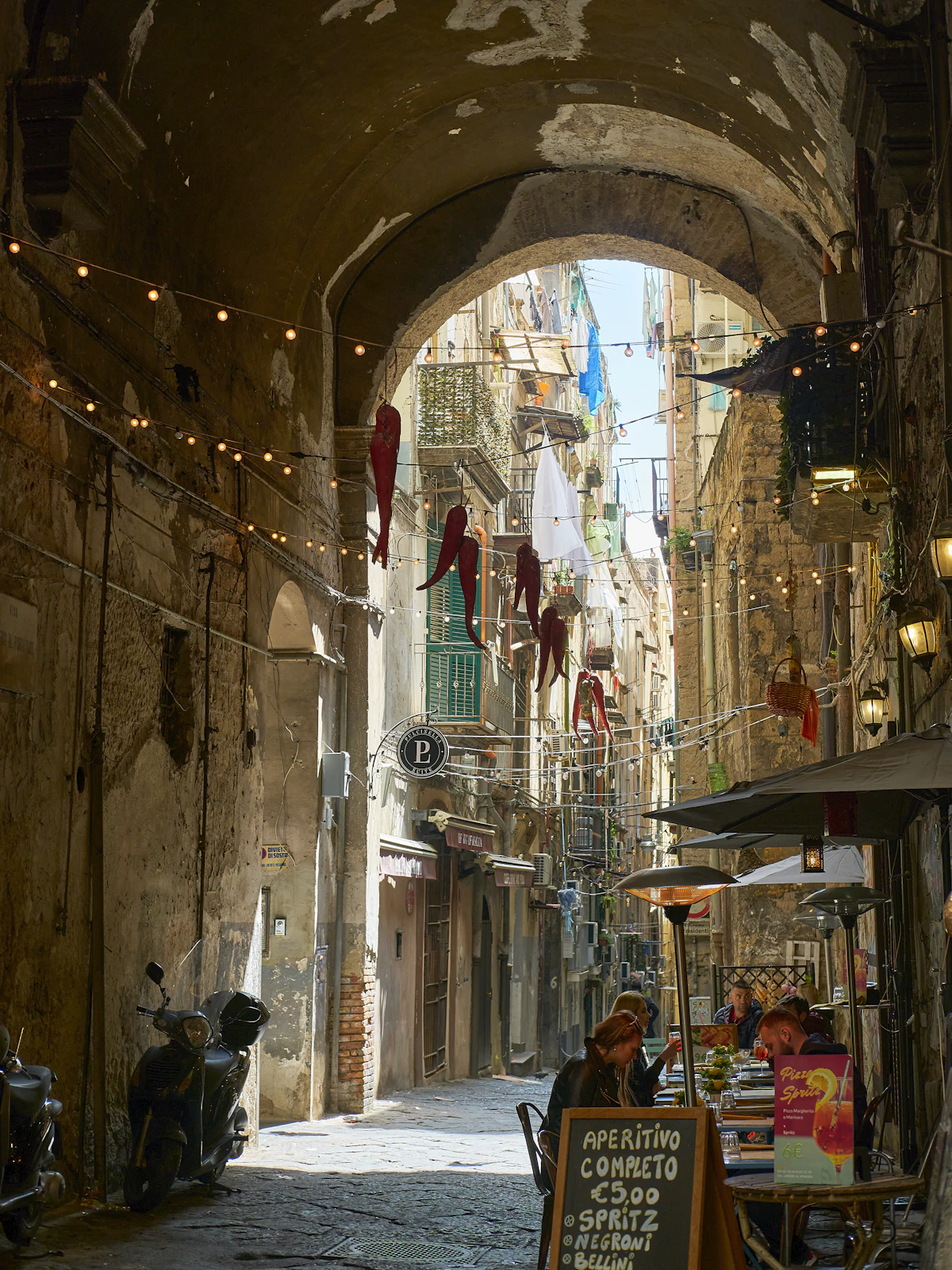 Uno stretto vicolo storico di Napoli con luci appese, portafortuna a forma di peperoncino e tavolini di caffè all'aperto (immagine rappresentativa di un vicolo del centro storico, non confermata come foto di Via San Gregorio Armeno stessa)