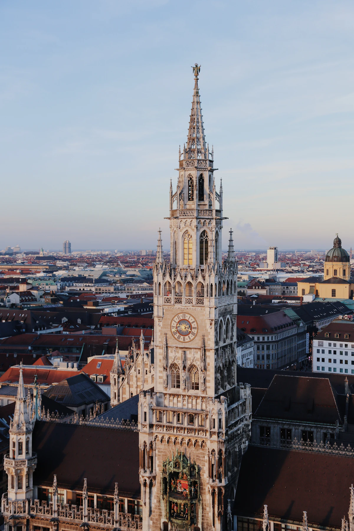 Die neugotische Spitze des Turms des Münchner Neuen Rathauses von oben gesehen, mit den Glockenspiel-Figuren an seiner Basis und der Stadtsilhouette dahinter